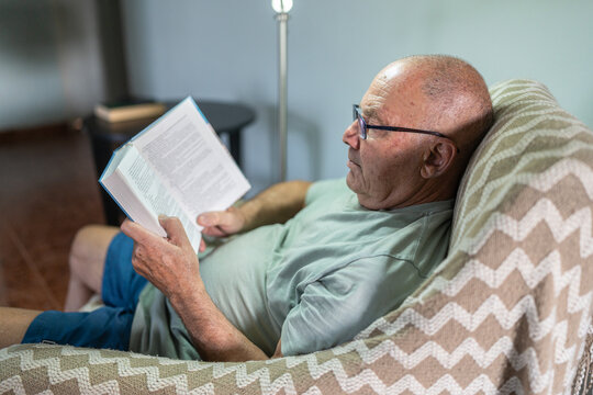 A senior man sits comfortably in a large chair, focused on reading a book. Soft light illuminates the space, creating a warm and inviting atmosphere perfect for enjoying literature.
