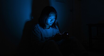 Young teenager using smartphone in dark room. Screen light illuminates his face with blue glow. Digital addiction and social media concept.