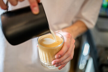 Barista pouring steamed milk into espresso to create latte art in a glass cup. Close-up of hands, heart-shaped foam design, specialty coffee preparation.