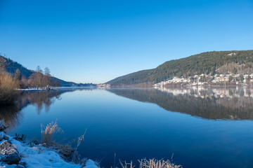 serene winter lakeside scene with snow-covered banks, blue water lake gerardmer, French Vosges, forested hills in distance, frozen landscape snow, still water, and bare trees, Seasonal beauty france