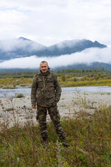 Naklejka premium A Caucasian man in camouflage clothing stands next to a dirty SUV in a grassy field. Mountains and clouds are visible in the background.