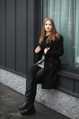A woman in a skirt and coat stands against a wall on the street.
