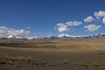 A landscape with snow-capped mountains under a clear blue sky. The foreground shows a flat, barren area with scattered clouds above it.