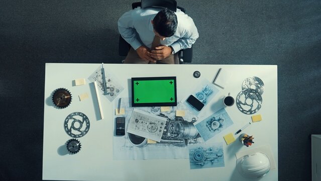 Top view of engineer looking at phone while working on tablet with green screen at meeting table with turbine engine or jet engine sketch design and various metal gear scatter around. Alimentation.
