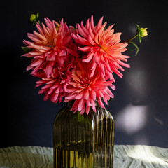 Bouquet of dahlias in green glass vase on dark background with sunlight.Vase of autumn flowers stands on table.Concept: greeting card, autumn holidays. Selective focus. Square