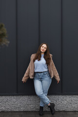 A girl in a winter parka, wearing blue jeans and a warm jacket. She has blond hair. She stands against a dark metal wall.