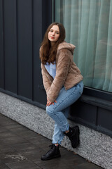 A girl in a winter parka, blue jeans, and a warm jacket. She has blond hair. She stands by a dark metal wall with a large window.