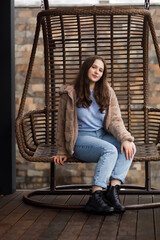 A girl in blue jeans and a warm jacket sits on a wicker bench on the terrace