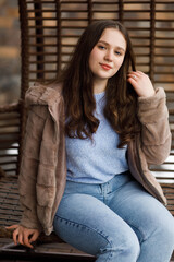 A girl in blue jeans and a warm jacket sits on a wicker bench on the terrace