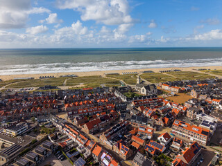 Fototapeta premium Aerial view of a coastal town with dense red roofed buildings, a tall steepled church, green spaces, wide sandy beach with huts, and ocean waves under a partly cloudy sky.