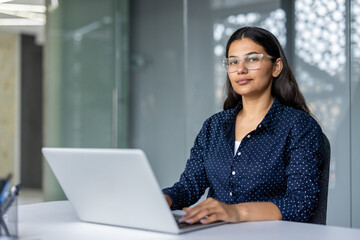 Indian businesswoman wearing glasses and a polka dot shirt sitting at a desk with a laptop,...