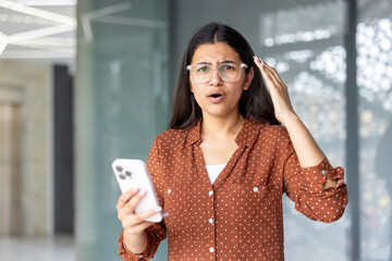 Woman with glasses holding a mobile phone and looking confused, shocked, and surprised, raising a...
