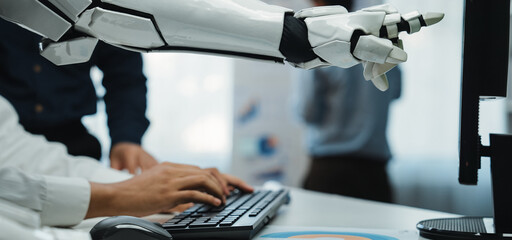 A close-up of a robotic arm assisting a person at a keyboard in a modern office, emphasizing collaboration between humans and machines for data tasks.Noogenesis