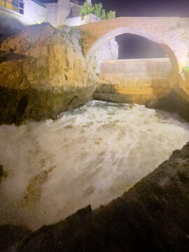 Puente de piedra sobre cauce rocoso con agua turbulenta iluminada de noche, bajo luz c&aacute;lida que resalta texturas.