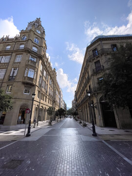 Avenida peatonal entre edificios cl&aacute;sicos con torre y balcones, pavimento geom&eacute;trico, farolas y cielo parcialmente nublado.