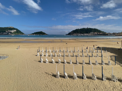 Playa de arena con filas de sombrillas cerradas frente a mar azul, con dos islas verdes y ciudad al fondo bajo cielo parcialmente nublado.