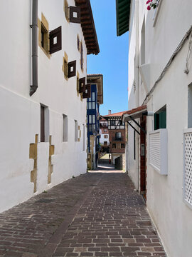 Callej&oacute;n estrecho entre fachadas blancas con detalles en madera y tonos azules, suelo de ladrillo y salida hacia edificios coloridos bajo cielo azul.