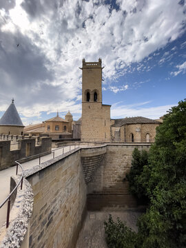 Torre rectangular de piedra con aberturas arqueadas, rodeada de edificios antiguos y patio inferior con vegetaci&oacute;n bajo cielo parcialmente nublado.