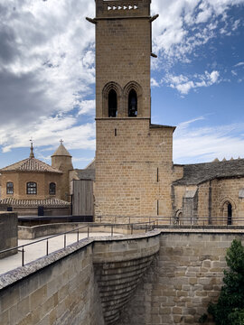 Torre rectangular de piedra con aberturas arqueadas, rodeada de edificios antiguos y muralla en primer plano bajo cielo parcialmente nublado.