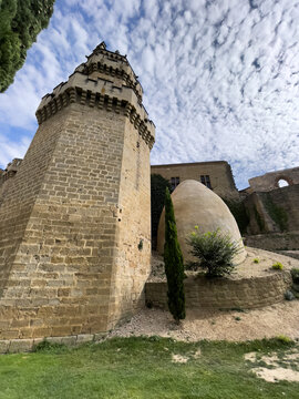 Torre de piedra con almenas junto a estructura abovedada, rodeadas de c&eacute;sped, arbustos y un cipr&eacute;s bajo cielo parcialmente nublado.