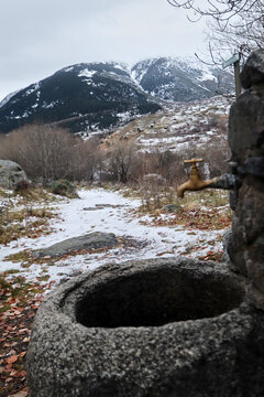 Pila de piedra con grifo met&aacute;lico frente a paisaje nevado, entre &aacute;rboles desnudos y monta&ntilde;as bajo cielo gris.
