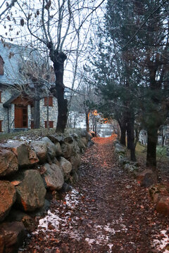 Sendero con hojas y nieve junto a un muro de piedra, conduciendo hacia una casa de monta&ntilde;a entre &aacute;rboles.