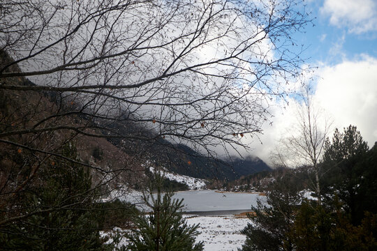 Lago parcialmente helado entre nieve y con&iacute;feras, enmarcado por ramas desnudas y monta&ntilde;as nubladas bajo un cielo azul con claros.