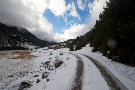 Camino nevado que curva entre con&iacute;feras y laderas oscuras, bajo un cielo azul con nubes que aportan luz al paisaje invernal.