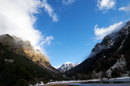 Valle de monta&ntilde;a con laderas nevadas y un lago que refleja el paisaje, bajo un cielo azul con nubes dispersas que iluminan la cumbre al fondo.