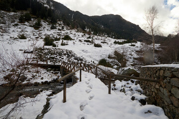Pequeño puente de madera sobre un arroyo en un entorno de montaña cubierto de nieve, con huellas que conducen hacia la estructura y árboles dispersos entre rocas y vegetación invernal, mientras las la © LIS