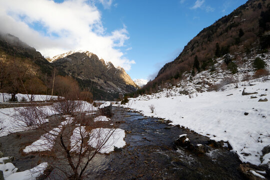 Valle de monta&ntilde;a con un r&iacute;o que avanza entre &aacute;rboles desnudos y zonas de nieve, mientras las laderas y cumbres se iluminan bajo un cielo parcialmente nublado, creando un contraste marcado entre la bla