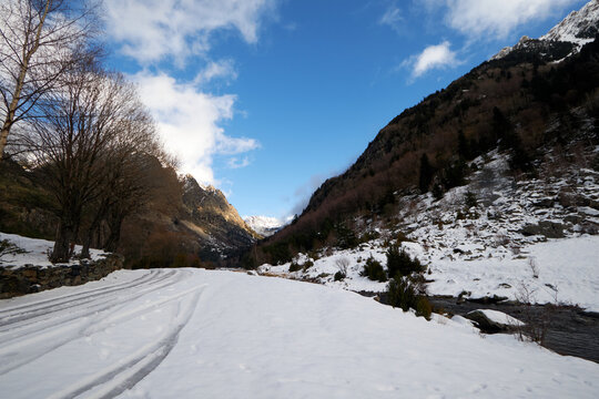 Valle de monta&ntilde;a cubierto de nieve con un arroyo estrecho que avanza entre &aacute;rboles desnudos y pendientes blancas, iluminado por un cielo claro con algunas nubes que realzan el contraste entre la nieve