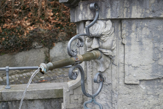 Fuente de piedra con rostro tallado, agua fluyendo por una boca met&aacute;lica y entorno oto&ntilde;al con escalones.