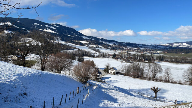 Paisaje invernal con colinas nevadas, &aacute;rboles desnudos y casas rurales, bajo cielo azul con nubes dispersas.