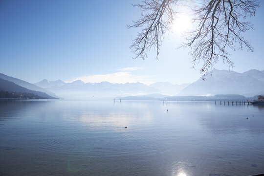 Lago sereno con monta&ntilde;as nevadas, reflejo brillante del sol y ramas desnudas enmarcando un muelle lejano.