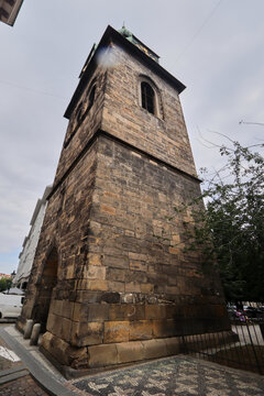 Torre alta de piedra con bloques envejecidos y aberturas arqueadas en la parte superior, rematada por un tejado met&aacute;lico verdoso con peque&ntilde;o pin&aacute;culo, junto a un paseo pavimentado y edificios urbanos 