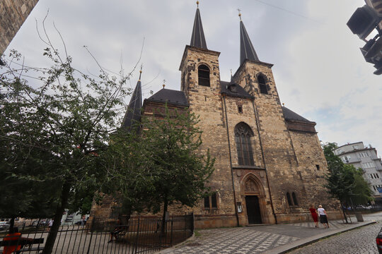 Iglesia hist&oacute;rica de piedra con dos torres altas rematadas en agujas, ventanales arqueados y un acceso central decorado, rodeada de un patio adoquinado con &aacute;rboles y algunas personas bajo un cielo par
