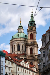 Fototapeta premium Iglesia barroca con torres de cúpulas verdes y reloj dorado, rodeada de edificios históricos de tejados rojos y cables de tranvía bajo un cielo parcialmente nublado.