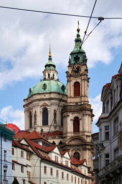 Iglesia barroca con torres de c&uacute;pulas verdes y reloj dorado, rodeada de edificios hist&oacute;ricos de tejados rojos y cables de tranv&iacute;a bajo un cielo parcialmente nublado.