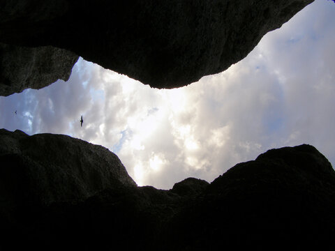 Vista ascendente desde una grieta rocosa hacia un cielo parcialmente nublado, con un ave volando en la distancia enmarcada por las paredes oscuras de la roca.