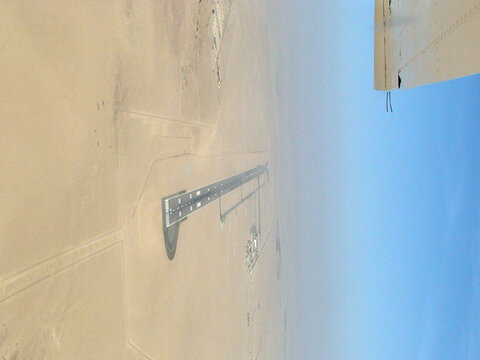 Vista a&eacute;rea de una larga pista de aterrizaje en medio de un desierto llano y arenoso, con algunas peque&ntilde;as estructuras alrededor y un cielo azul despejado sobre el paisaje &aacute;rido.
