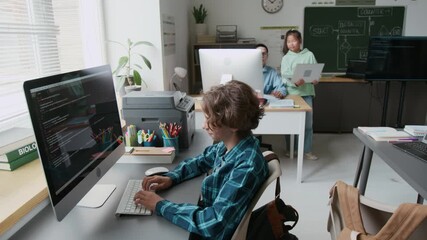 Smart boy typing code on computer while teacher working with other student during programming lesson in school - Powered by Adobe