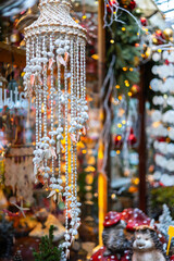 A close-up of a hanging wind chime made of shells and beads at a market stall. Christmas lights and other decorations are in the blurred background.