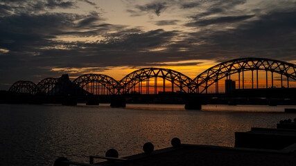 Obraz premium Extremely beautiful and peaceful scene of the Iron Bridge (Dzelzceļa tilts) over the Daugava River at sunset, with its round iron arches standing out against the vibrant orange and blue evening sky.