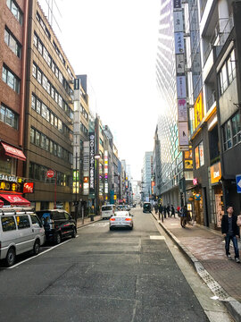Calle urbana japonesa con tr&aacute;fico moderado, r&oacute;tulos en edificios altos, comercios a pie de calle y peatones en aceras limpias. Escena din&aacute;mica de ciudad.