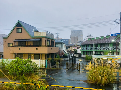 Barrio residencial urbano en d&iacute;a lluvioso, casas y edificios bajos, carretera mojada, vegetaci&oacute;n en primer plano y ambiente tranquilo. Escena cotidiana de ciudad.