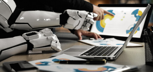 A futuristic workspace showcasing a human collaborating with a robotic arm, analyzing digital designs and creative concepts on a laptop, emphasizing innovation and teamwork.Noogenesis