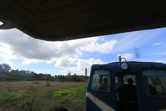 Fotograf&iacute;a de un vag&oacute;n de tren azul en primer plano, con pasajeros visibles en su interior, atravesando un paisaje rural de campos verdes y cielo parcialmente nublado. La escena transmite una sensaci&oacute;