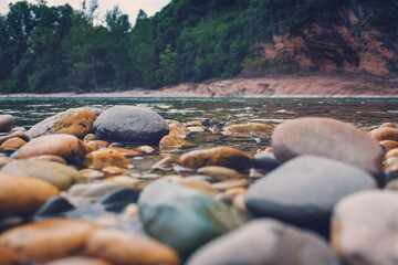 Smooth river stones in various colors are partially submerged in clear water, surrounded by lush greenery and rocky cliffs, creating a serene natural landscape