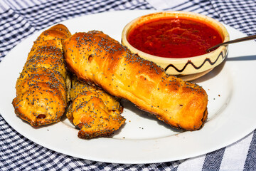 Sausages baked in dough sprinkled with salt and poppy seeds and bowl with tomatoes sauce in a rustic composition. Sausages rolls, delicious homemade pastries.